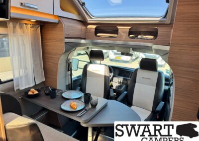 Camper van dining area with a table set for a meal: two plates, croissant, eggs, cups, and cutlery, wood cabinets and a skylight above; front seats visible.