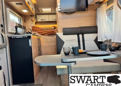 Interior of a camper van: kitchen on the left with fridge and stove, a two-person dinette with a table set for two, bed and storage at the rear, a wall-mounted TV, and windows with white curtains; SWART CAMPERS logo bottom right.