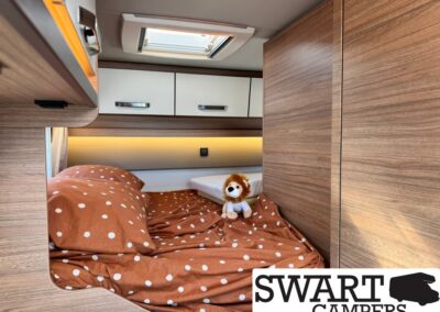 Interior of a camper bedroom with polka-dot brown bedding and a small lion plush on the bed, wood cabinets above and beside.