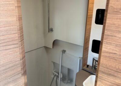 Compact camper shower with vertical chrome rail, handheld shower, and white molded walls; sink and wood cabinet visible nearby.
