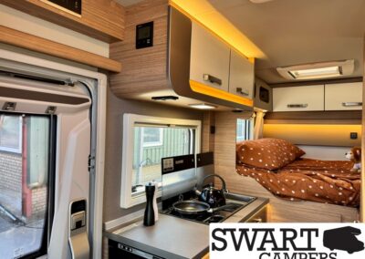 Interior of a compact campervan: small kitchen with stove and kettle, overhead cabinets, and a bed with brown polka-dot bedding.