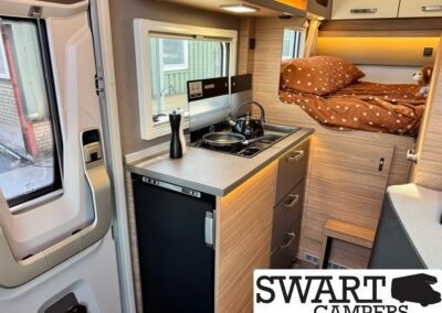 Interior of a compact camper van: kitchenette with two-burner stove, kettle, window, and brown polka-dot bed area in the back; warm wood cabinets and drawers.