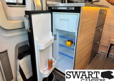 Open mini fridge in a camper kitchen with a yellow bell pepper on the shelf and a can in the door pocket, wooden cabinetry nearby.