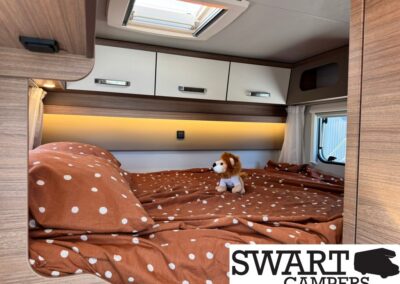 Cozy camper van bed with brown polka-dot duvet, a plush lion, white overhead cabinets, and a skylight.