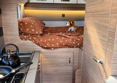 Interior of a camper: loft bed with brown polka-dot duvet, a small lion plush toy, and a compact kitchenette with kettle on the stove.
