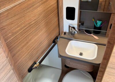 Compact camper bathroom with a small sink, round toilet, and a shower tray, all surrounded by wood-paneled cabinets.
