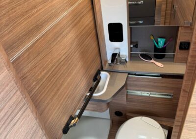 Compact camper bathroom with wood veneer cabinets, a small shower base, fold‑down sink, mirror cabinet, and a toilet.