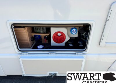 Exterior service bay of a camper with a red circular cap and a blue water-fill cap inside an open compartment.