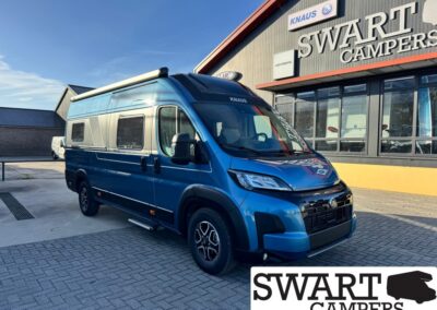 Blue camper van with a raised roof parked at a Swart Campers showroom, with the dealership name visible on the building behind it.