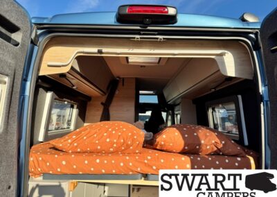 Interior of a camper van with a raised bed in orange polka-dot bedding and wooden cabinetry inside a compact living space