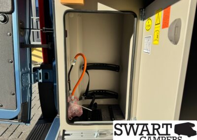 Open storage compartment of a camper van with an orange hose, black straps, and a small wrapped item inside.