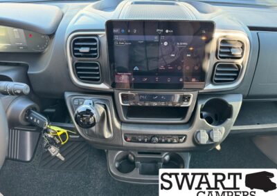 Left-hand-drive van dashboard featuring a large central touchscreen, climate controls, gear selector, cup holders, and keys in the ignition; SWART CAMPERS logo appears in the bottom-right corner.