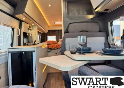 Interior of a modern camper van with a kitchenette on the left, a small table set for two, and a bed visible toward the rear; warm lighting.