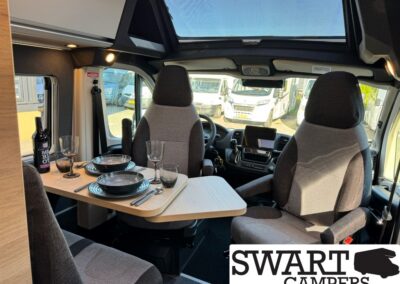 Interior of a camper van with a wooden table set for two, plates, wine glasses, and a bottle, front seats and a sunroof visible outside the cab area.