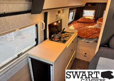 Compact campervan kitchenette with a two-burner stove and sink on a light wood counter, next to a windowed wall and sliding storage drawers, leading to a cozy bed area with orange polka-dot bedding and a teddy bear.