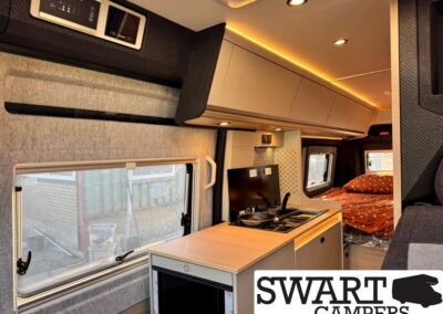Interior of a camper van with a wooden countertop kitchen, small stove, sink, and overhead cabinets, a window on the left, and a cozy bed with orange bedding toward the back, lit by warm LED lights.
