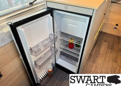 Open mini fridge in a camper kitchen, two transparent door shelves, a can on the door, and two peppers on the main shelf.