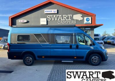 Blue camper van parked in front of a Swart Campers building with logo signs on the facade and a graphic panel on the side against a clear sky.