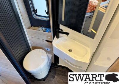Compact camper bathroom with a white oval toilet beside a curved white sink and a large mirror, wood cabinetry, and a small window by the door.