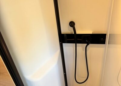 Interior of a camper shower: white walls, a black wall panel with a handheld shower hose, and a wooden top shelf.