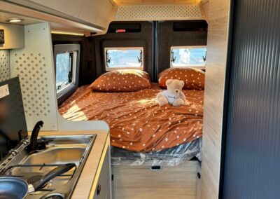 Compact camper-van interior: kitchenette on the left and a rear bed with orange polka-dot bedding and a teddy bear.