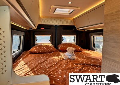 Cozy camper-van bedroom with orange polka-dot bedding, two pillows, and a small teddy bear on the bed, framed by side windows and a skylight.