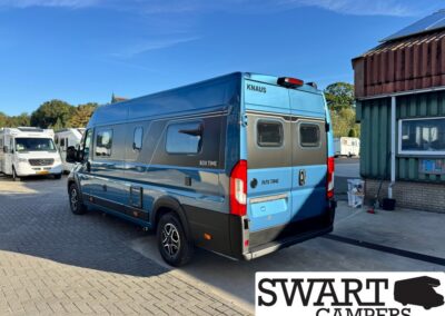 Blue Knaus Box Time camper van parked in a lot with other motorhomes nearby and a green wooden building on the right; Swart Campers logo visible in the bottom-right corner.