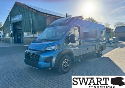Blue Knaus camper van parked in a sunny lot beside a workshop labeled 'Garage Swart'.