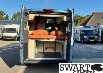 Blue camper van with rear doors open showing a raised bed with orange bedding and storage below, in a dealership lot with other campers nearby.