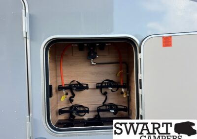 Open exterior storage bay of a camper showing a built-in bike rack and orange hoses inside; Swart Campers logo bottom right.