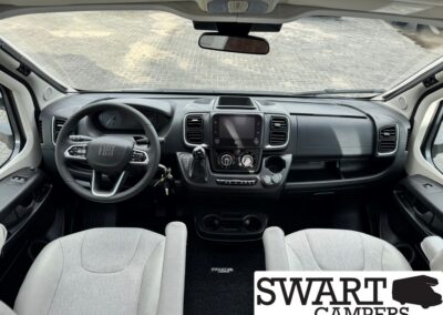 Interior view of a Fiat camper van cockpit: steering wheel, central touchscreen, gear shifter, and climate controls seen from the driver’s seat, with gray seats and dashboard.