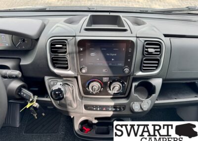 Dashboard view of a modern vehicle interior with a central touchscreen, two air vents, and climate controls below, plus gear selector and cup holders nearby. SWART CAMPERS logo in the bottom-right corner.