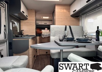 Camper van interior with a booth-style dining area set for two, wine bottle and glassware on the table, and a kitchen to the left with light wood cabinets; bed visible in the rear storage area.