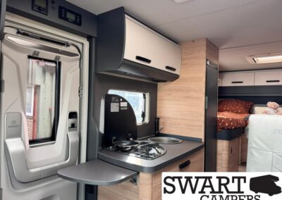 Interior of a modern camper van with a compact kitchen: two-burner stove, sink, fold-out counter, wooden cabinets, and a sleeping area in the back.