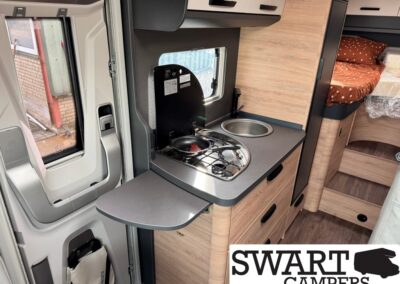 Camper van kitchenette with a two-burner stove, small sink, and a fold-out gray countertop. Wooden cabinets line the area, with a bed visible in the background.