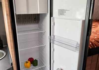 Compact camper fridge with peppers (yellow, red, green) on lower shelf and a can on door shelf; empty top shelves in a small kitchen area.