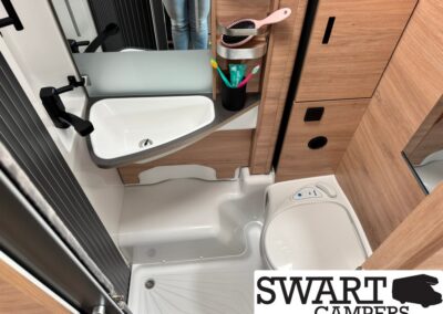 Compact camper bathroom with white sink, black faucet, and wooden cabinetry; toothbrushes and grooming brushes in a holder above the counter; small mirror and storage shelves visible in a narrow space. A portable toilet sits on the floor to the right; light-colored shower base is at lower left; Swart Campers logo partially visible in the corner.