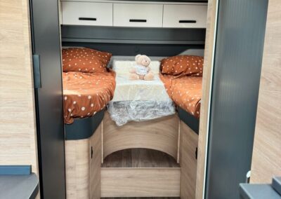Small camper bedroom with two single bunks on either side, orange polka-dot bedding, and a teddy bear at the center on a plastic-wrapped mattress. Overhead white cabinets and a skylight above. Wood-toned interior and steps/curtain area at the foot of the bed.