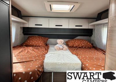 Interior of a camper bedroom with two single beds in brown polka-dot bedding, a stuffed bear between them, and white overhead cabinets.