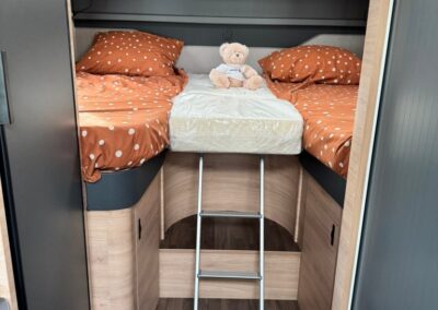 Camper bunk bed area with orange polka-dot bedding, a teddy bear, and a small metal ladder leading up to a mattress under overhead cabinets.