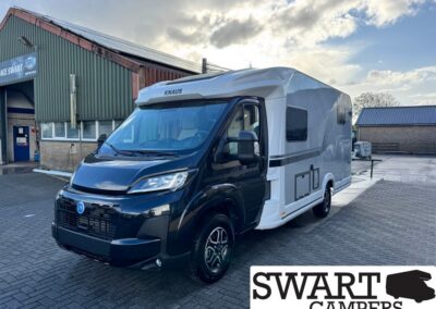 White and black KNAUS camper van parked in a lot beside a green industrial building with SWART CAMPERS logo visible in the corner, front view.