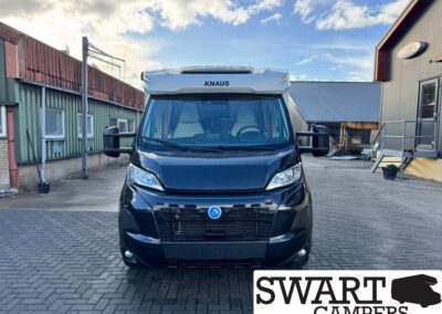 Front view of a black KNAUS camper van parked in an industrial yard, with blue sky above.
