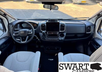 Front cabin view of a Fiat camper van showing the steering wheel, center touchscreen, gear shifter, and two light-gray front seats.