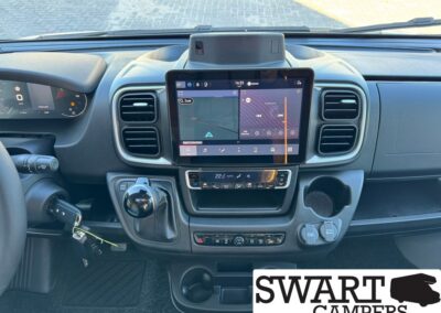 Dashboard of a camper van with a large center touchscreen, climate controls, and gear lever; keys in ignition and Swart Campers logo in the corner