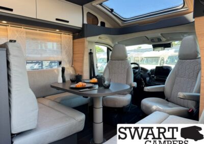 Interior of a camper van with a round table, gray seating, and pastries on plates under a skylight in front of the cabin area. SWART CAMPERS logo visible at bottom right.