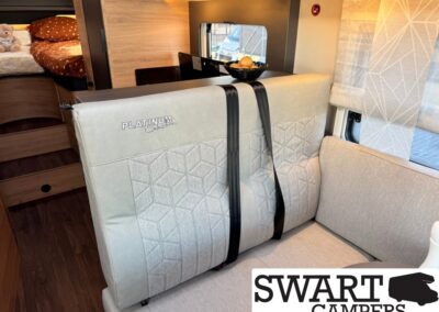 Camper van lounge area with light gray seating, black seat belts, wood paneling, and a fruit basket on the counter; bed with a teddy bear visible in the back.