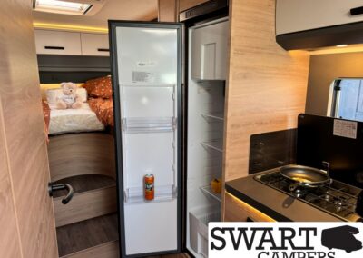 Open refrigerator in a compact camper van kitchen with a stove to the right and a bed with an orange duvet in the background (teddy bear on the bed).