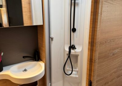 Compact caravan bathroom featuring a tall white shower enclosure with a handheld shower, and a white sink with rounded basin on a wood-paneled wall. Swart Campers logo visible in corner.