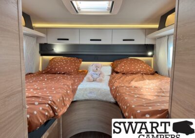 Twin bed setup in a camper interior with orange polka-dot duvet, a stuffed bear between pillows, and overhead cabinets above the headboard.