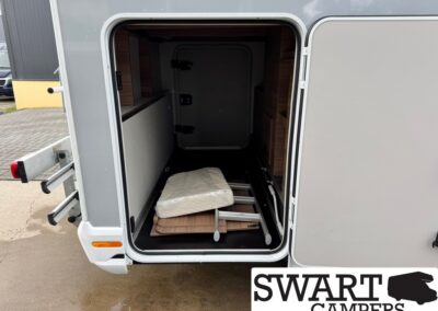 Open rear storage compartment of a campervan with disassembled furniture pieces (wood panels, metal frame) and a covered mattress on the floor inside.